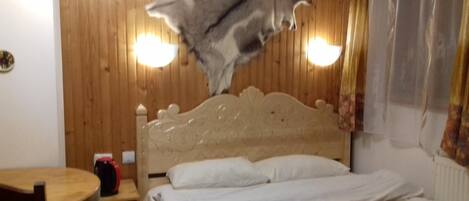Una habitación con cabecera de madera, cama con ropa de cama blanca, alfombra estampada y una mesa con un objeto rojo encima.