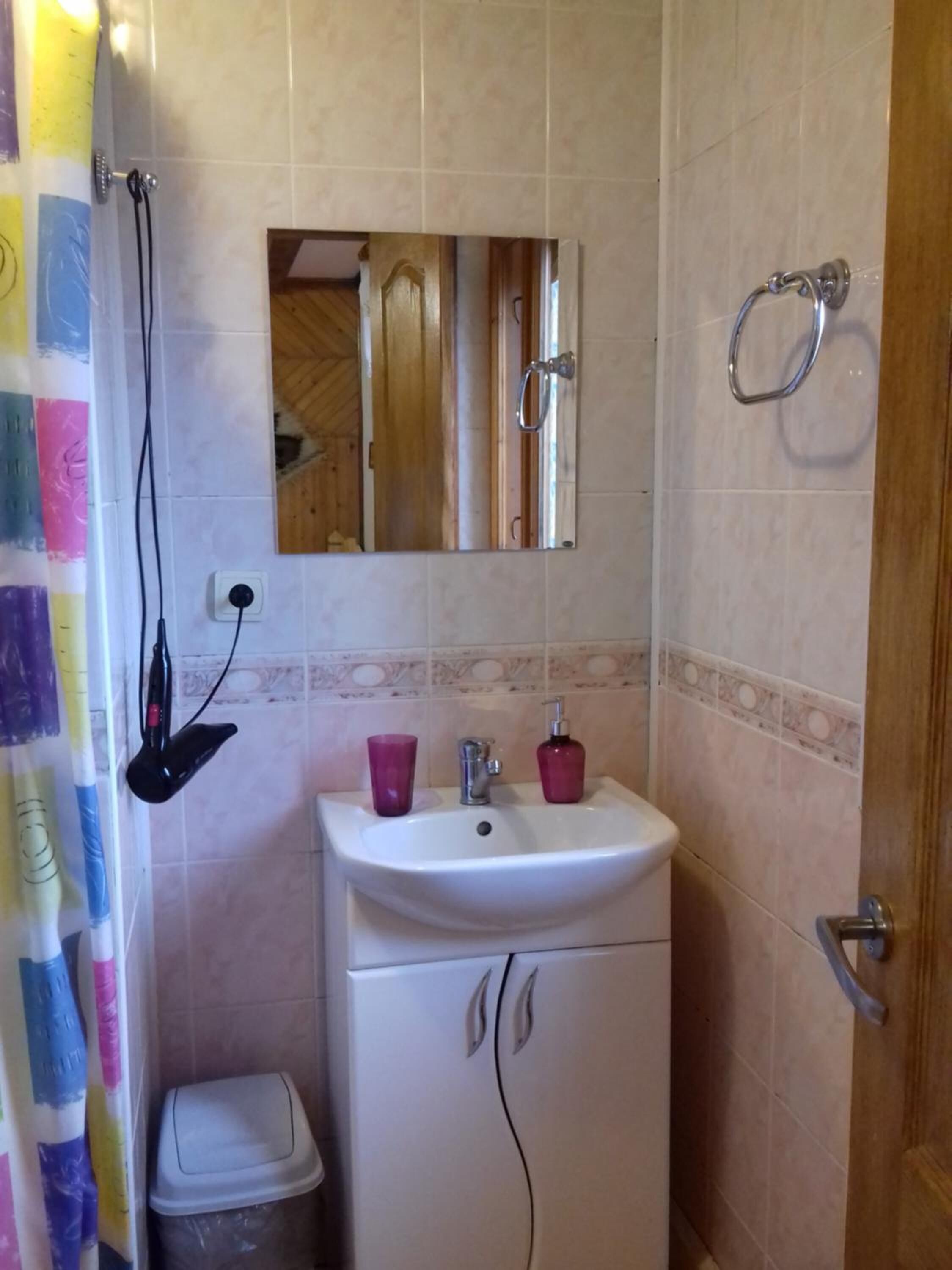 A small bathroom with a white sink, mirror, and shower. There is a trash bin and a door to the right.