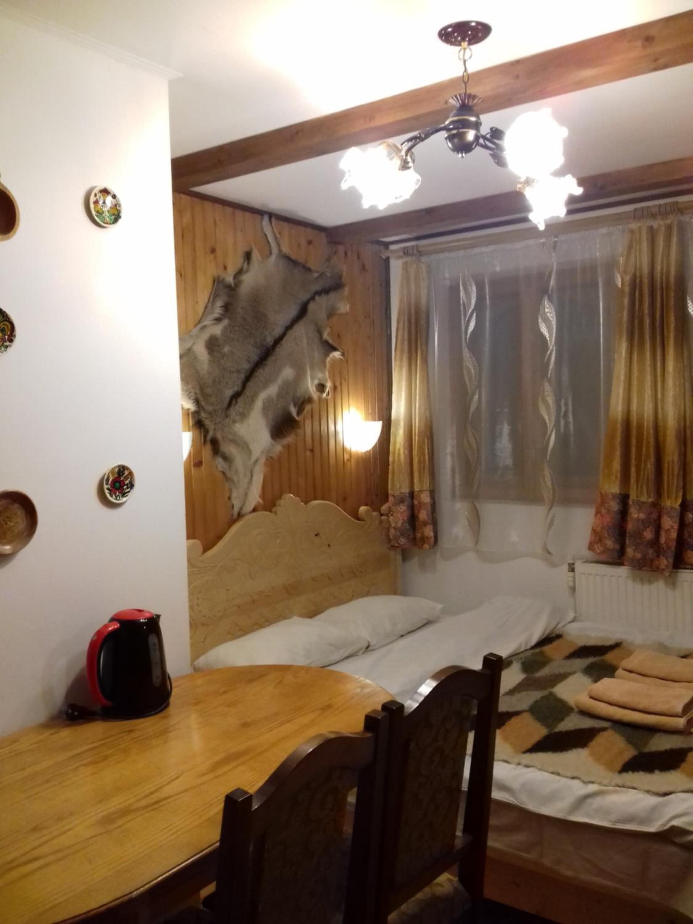 A room with a wooden headboard, a bed with a patterned blanket, a wooden table with a red kettle, and a fur wall piece.