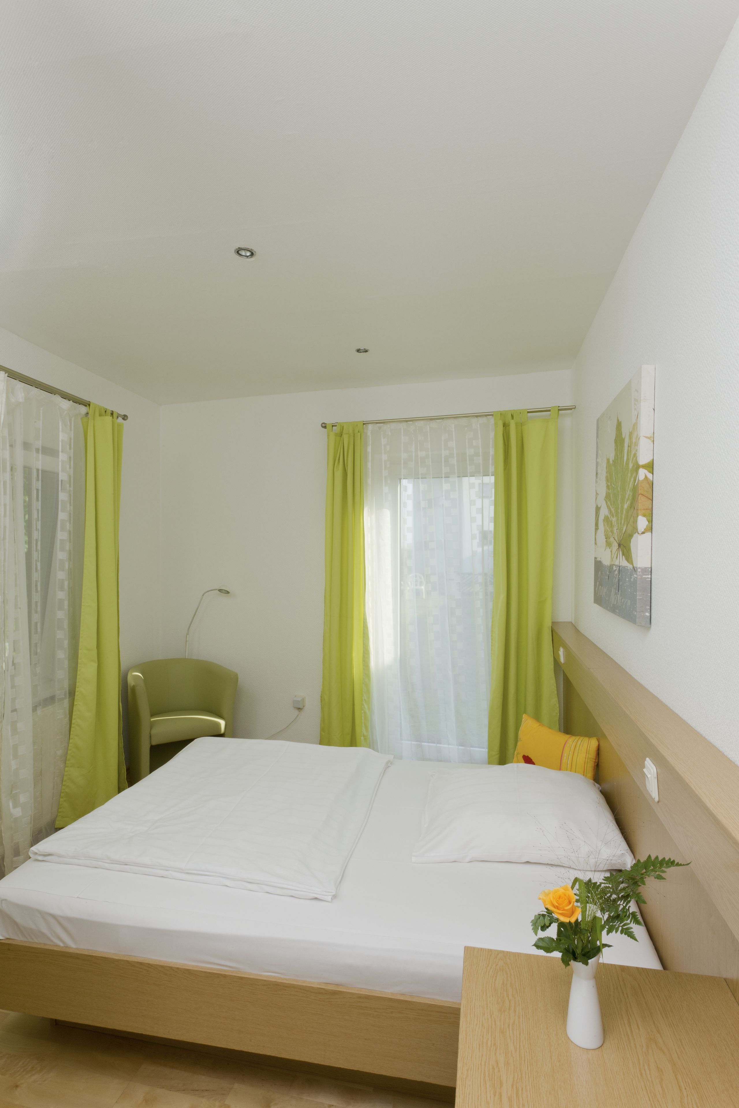 A neatly made bed with a white mattress and a wooden headboard. There is a green chair and a small table with a vase of flowers.