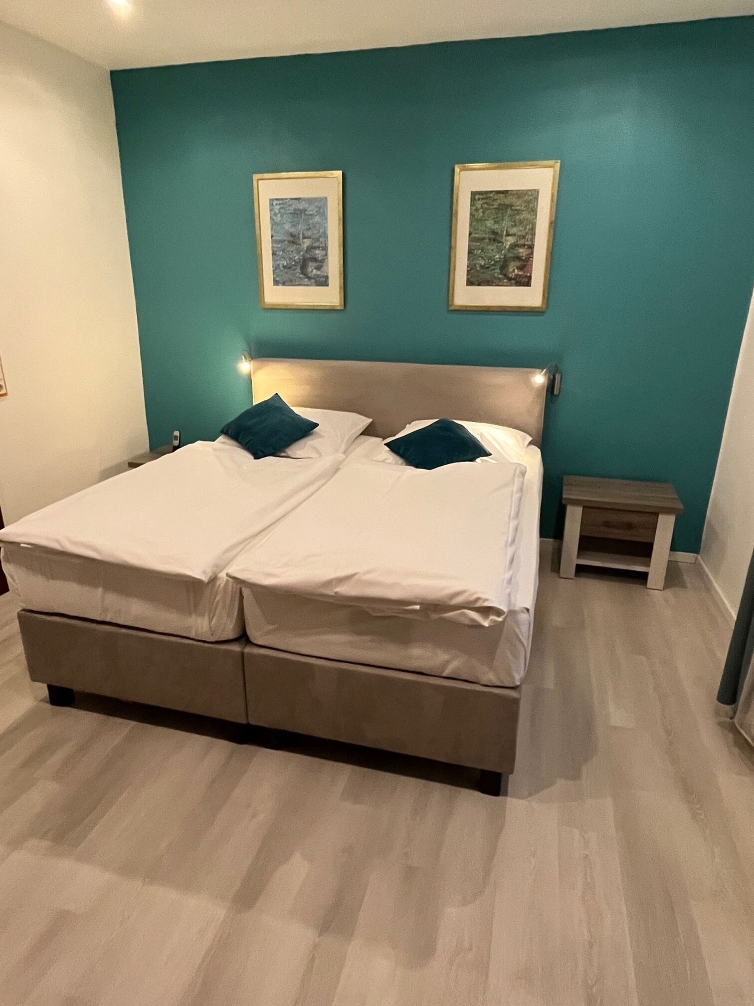 A double bed with white linens and two pillows, a wooden nightstand, and two framed pictures on teal walls.
