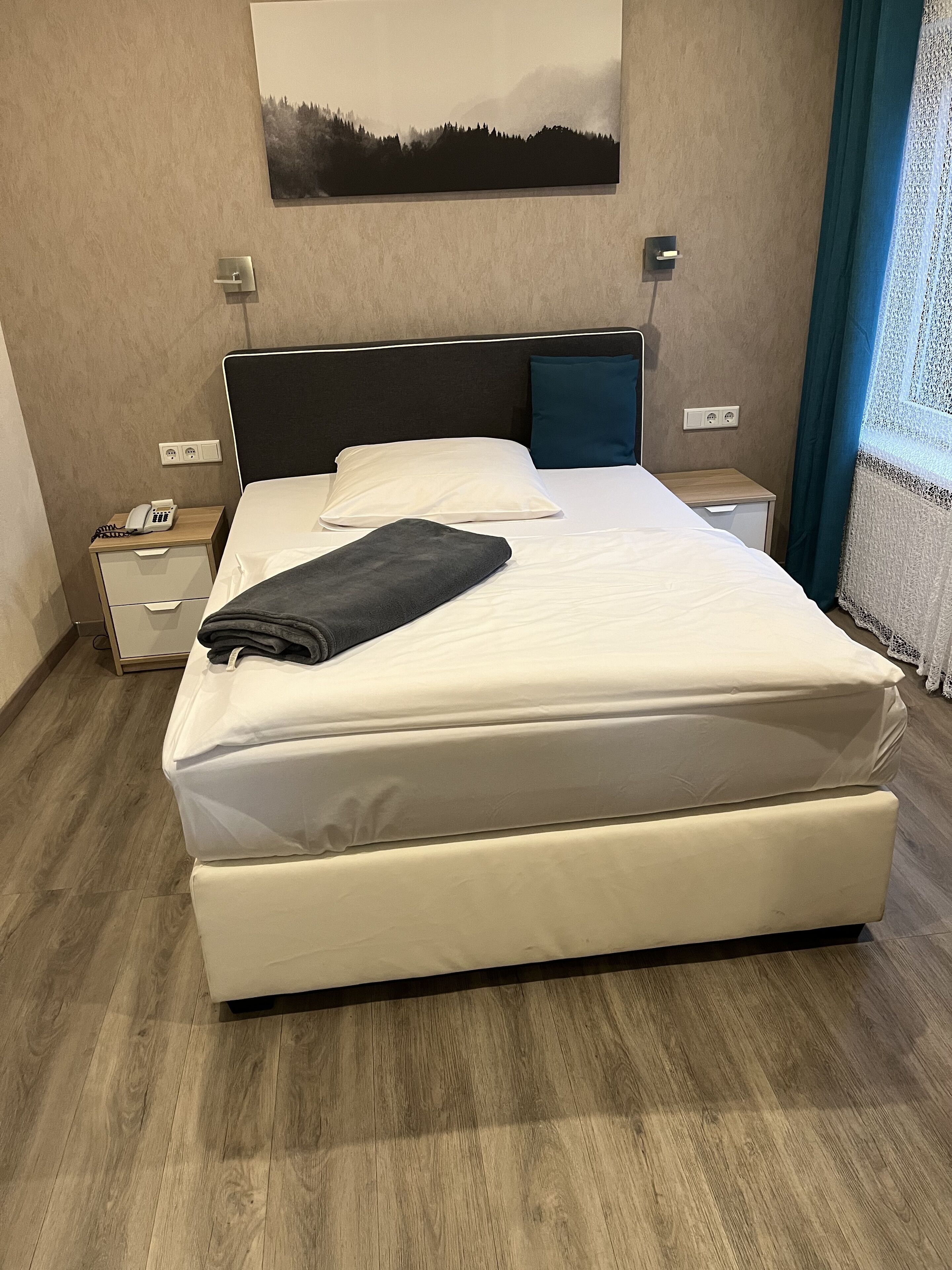 Una cama bien hecha con un colchón blanco y una manta oscura, una mesita de noche con un teléfono y una lámpara.
