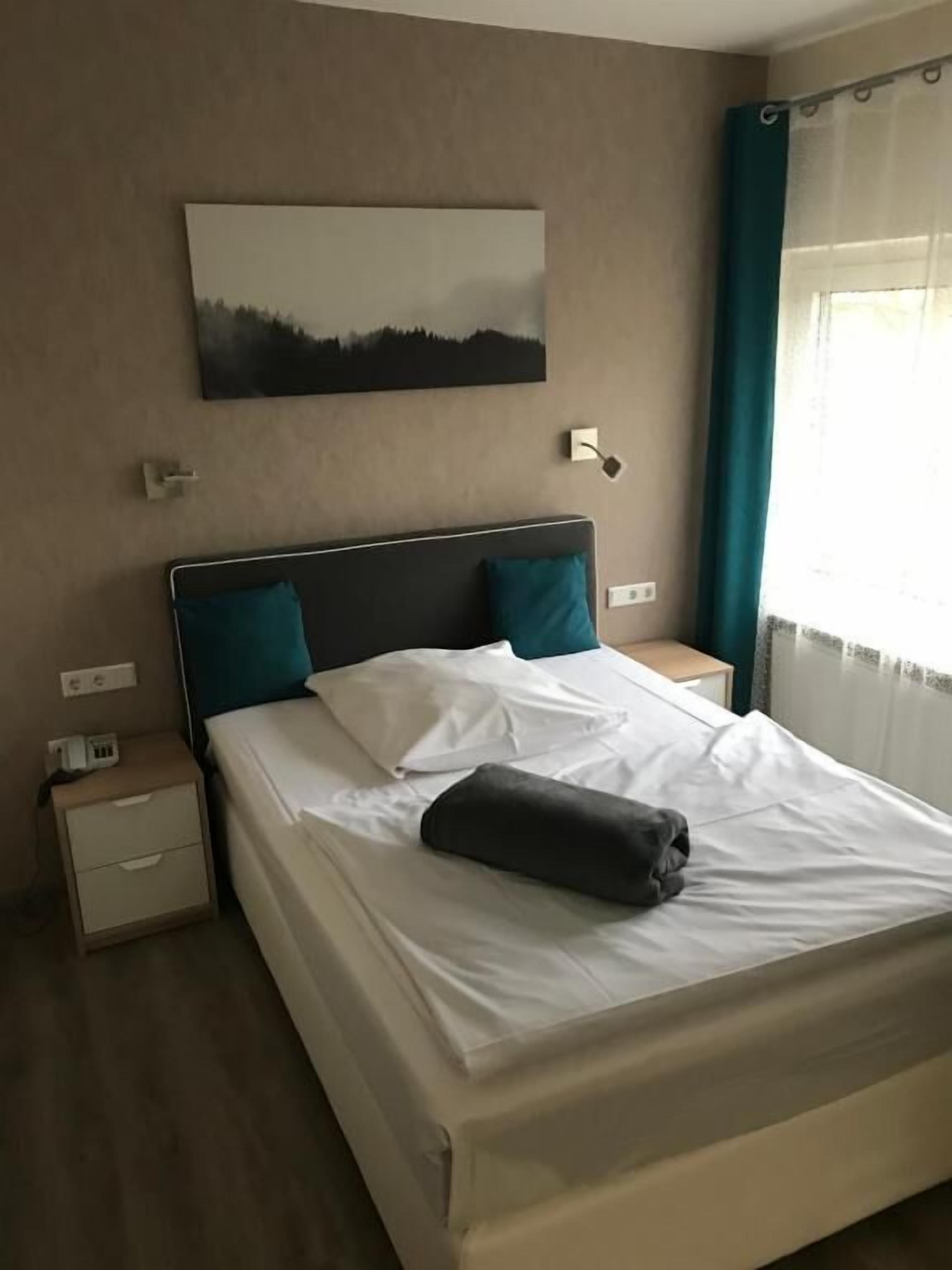 Una cama bien tendida con sábanas blancas y dos almohadas color aguamarina, una mesita de noche con una lámpara, y un cuadro de un paisaje en la pared.