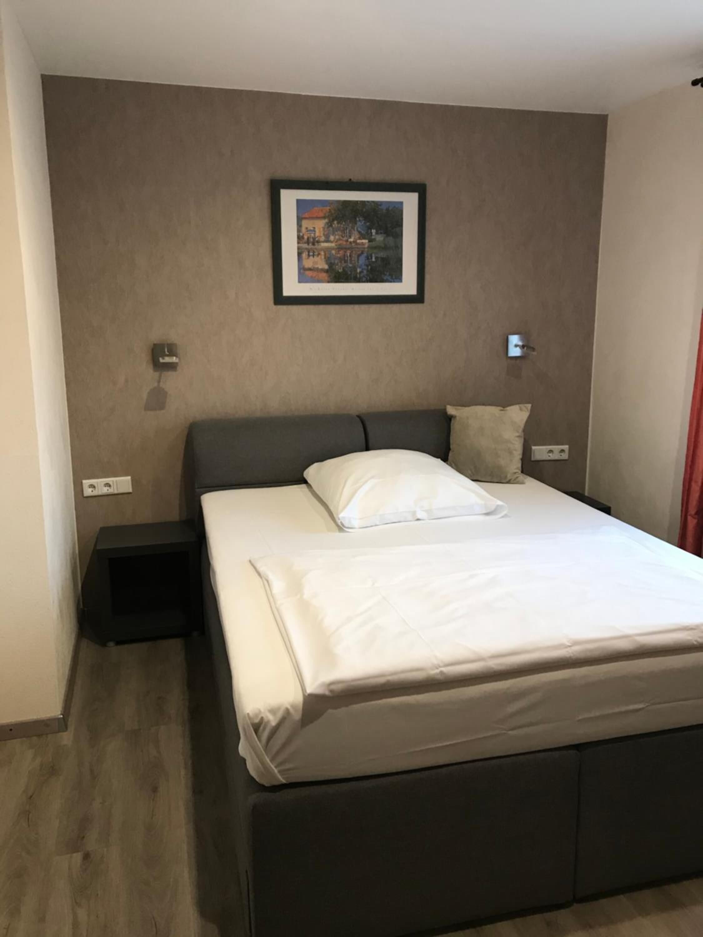Un lit bien fait, avec du linge de lit blanc, dans une chambre moderne avec un mur marron et un tableau encadré au-dessus.