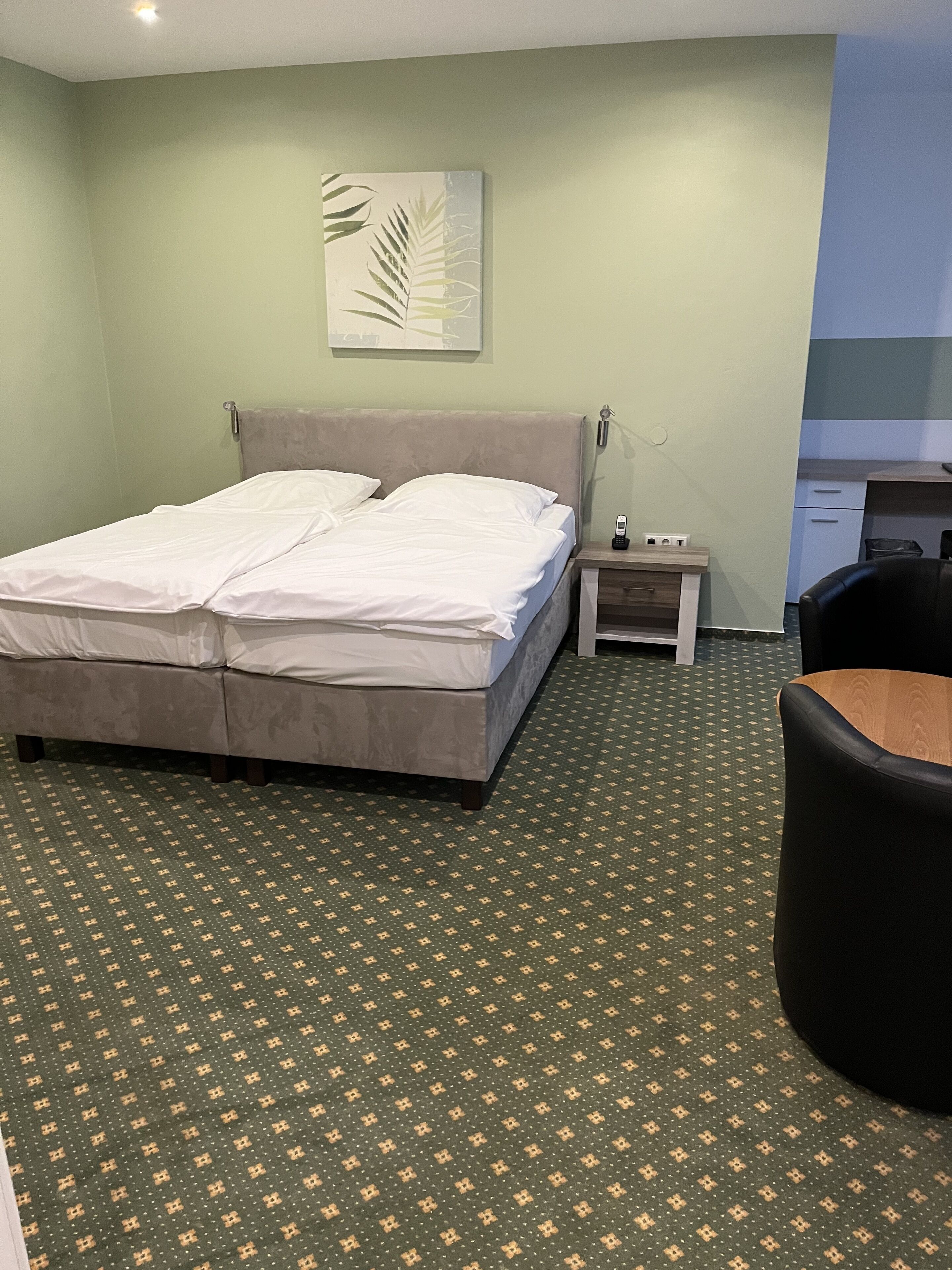 Et hotelværelse med en dobbeltseng, et lille natbord med telefon, en stol og et grønt mønstret tæppe.