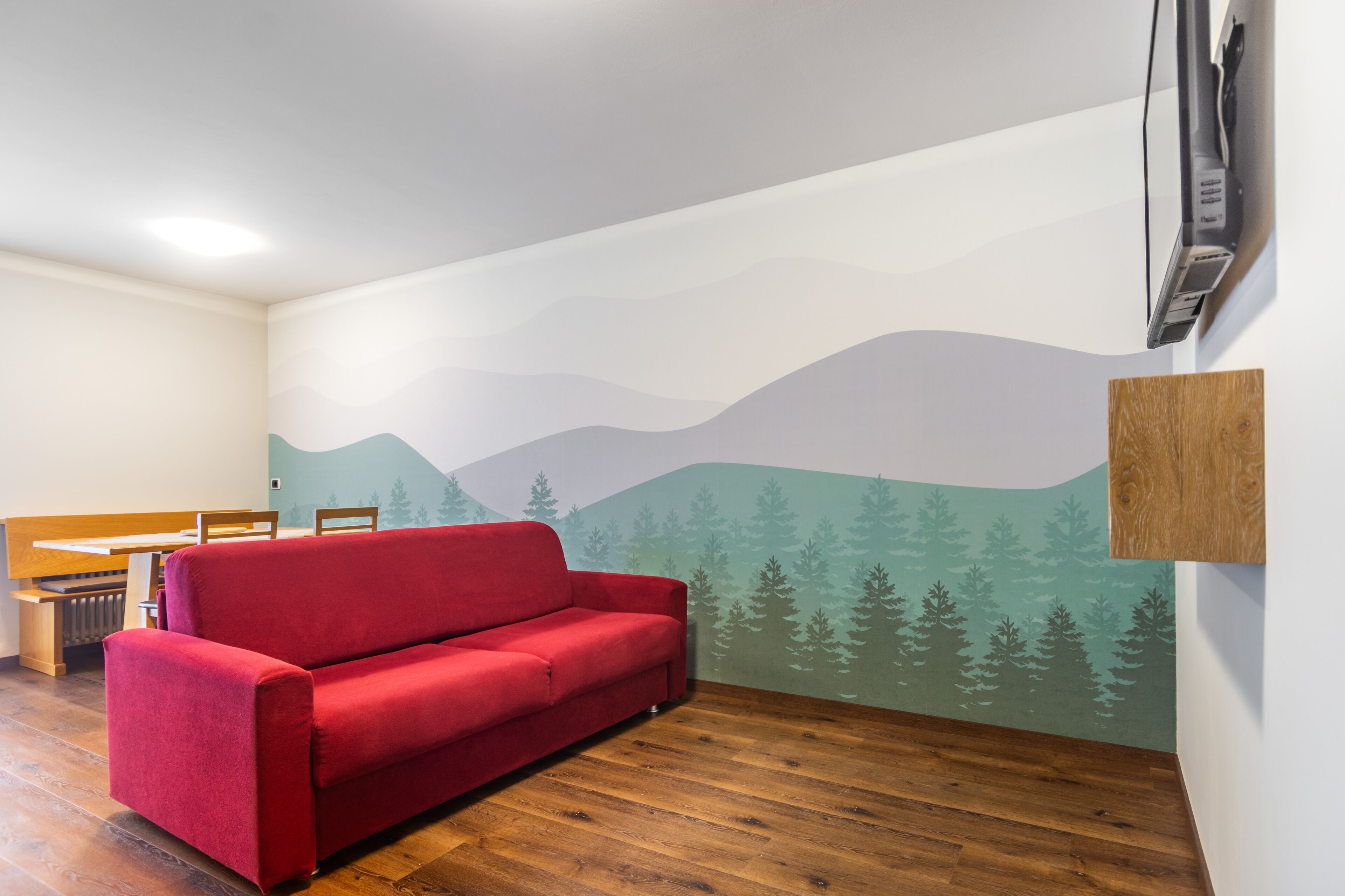 A room with a red sofa, wooden flooring, a mural of mountains and trees, a wooden bench, and a flat-screen TV mounted on the wall.