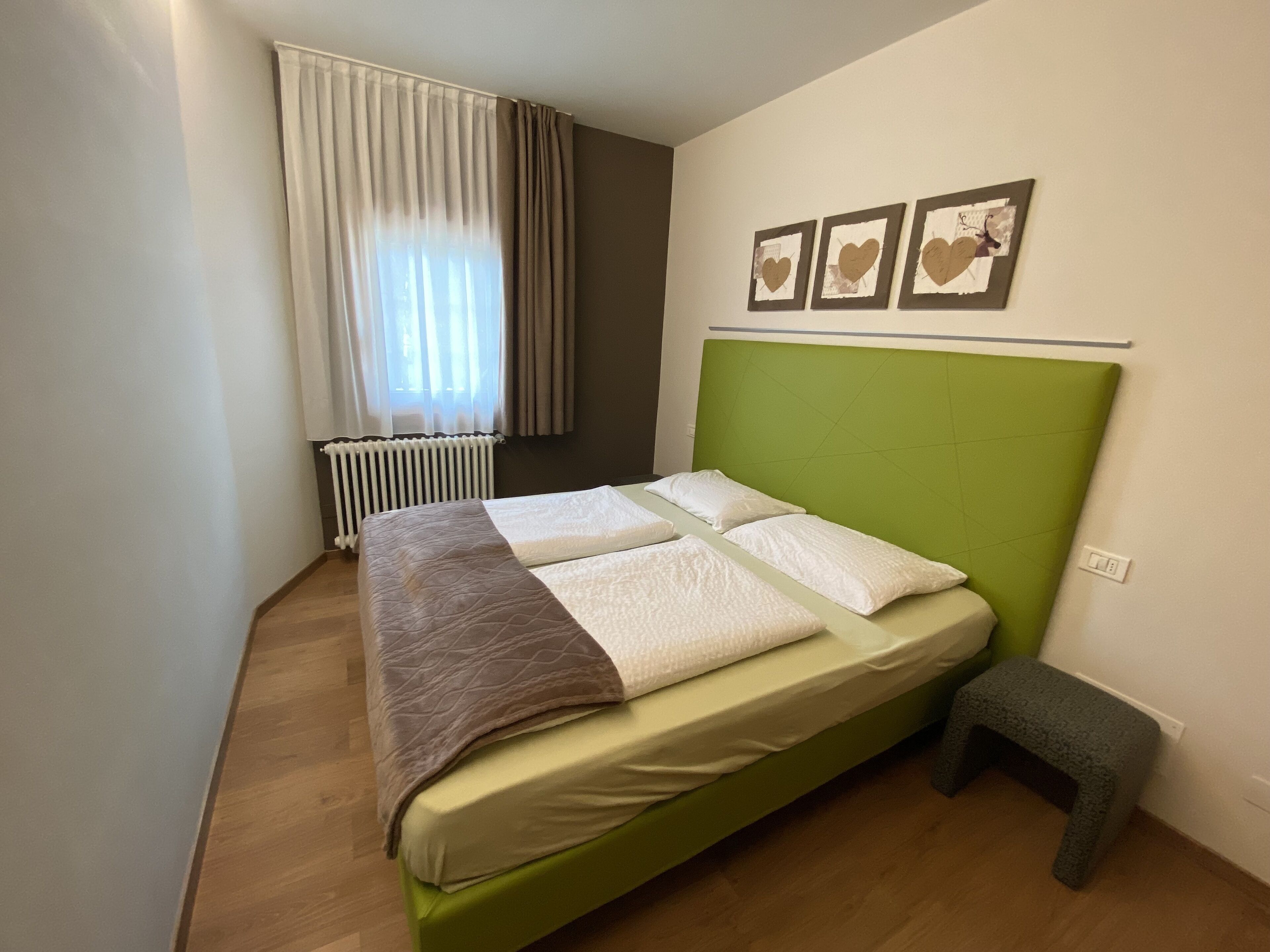 Een net opgemaakt bed met een groen hoofdeinde, een nachtkastje en een radiator in de hoek.
