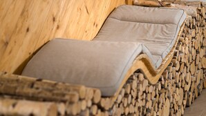 A wooden log cabin with a cozy lounge area featuring a cushioned bench and a stack of firewood.