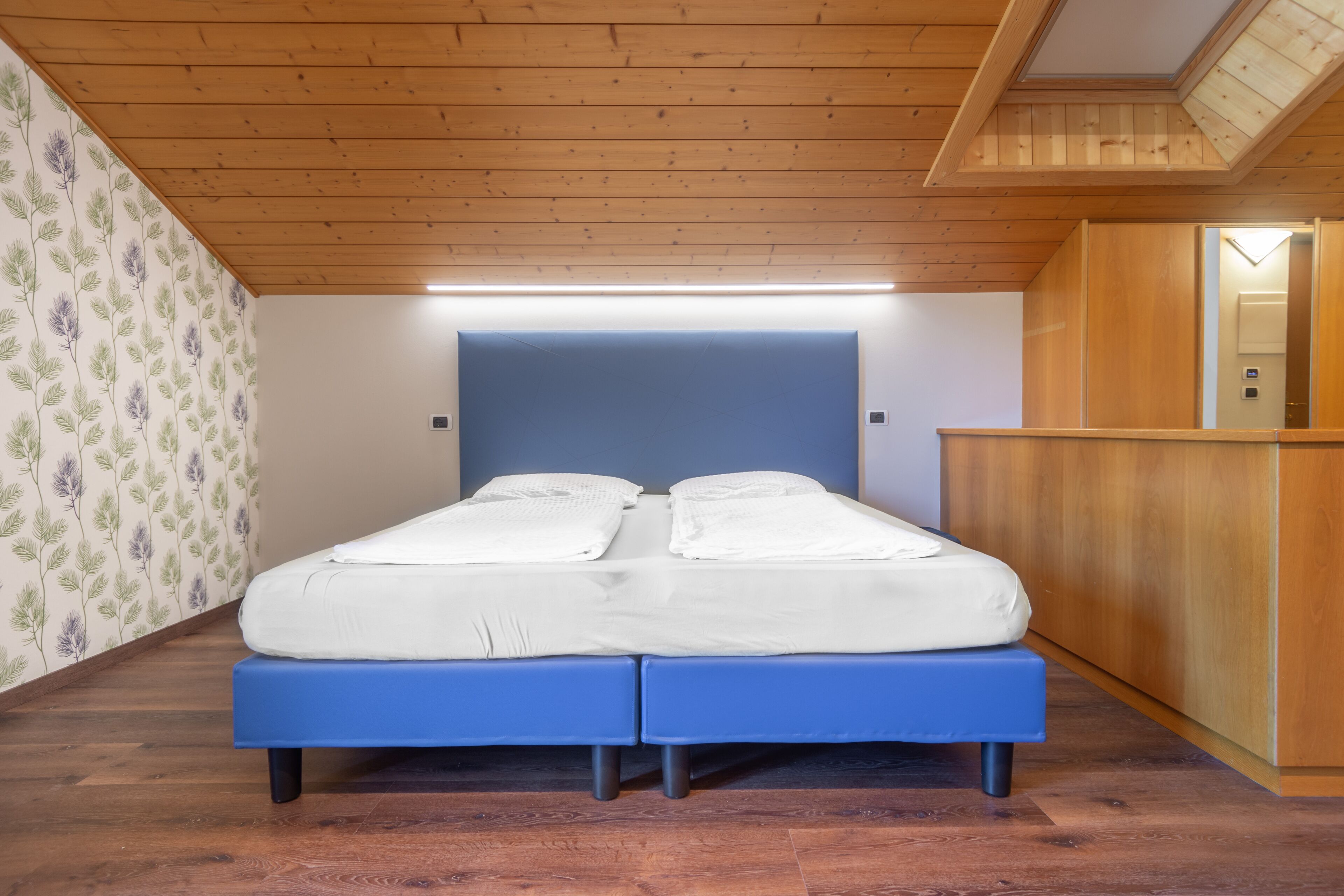 A bedroom with a wooden ceiling, a bed with a blue headboard, and a wooden wardrobe.