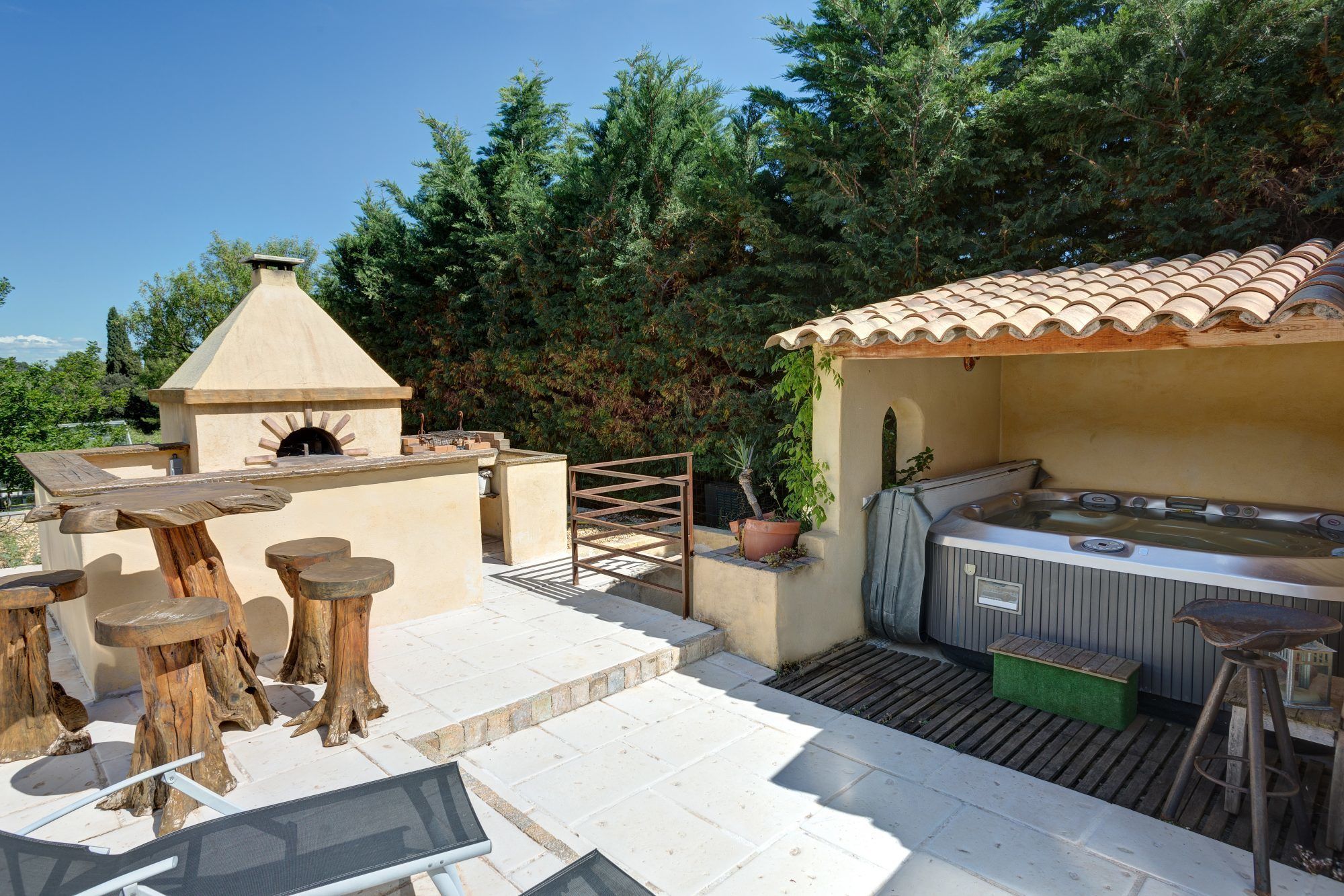 An outdoor patio with a stone table, wooden stools, a hot tub, and a pizza oven.
