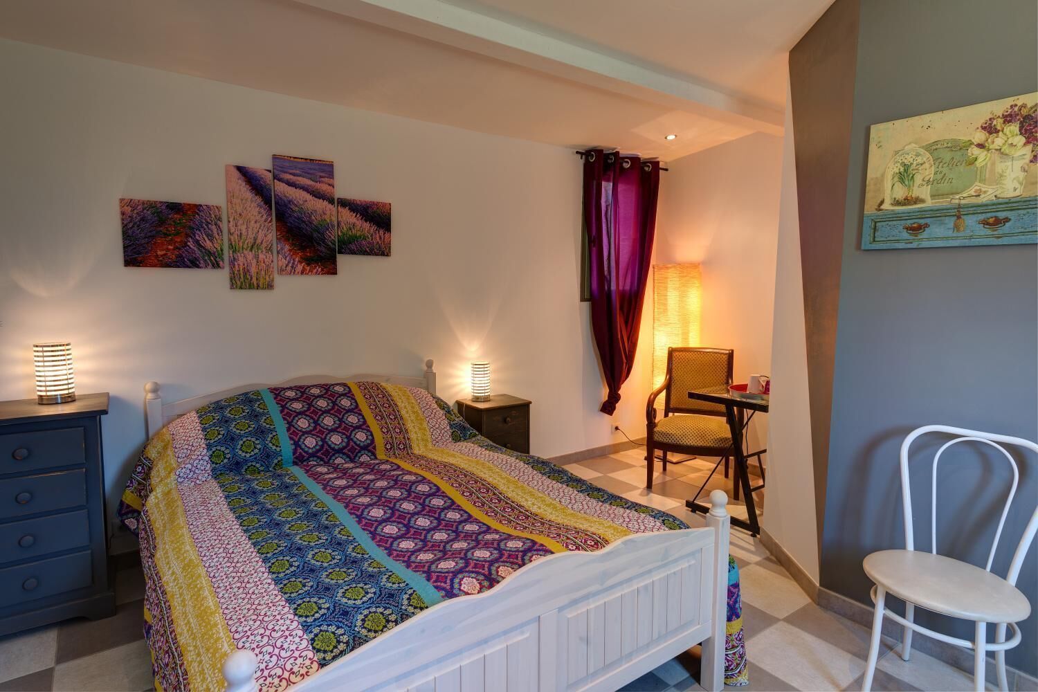 A bedroom with a bed, two bedside lamps, a chair, a small table, and a wall with three paintings.