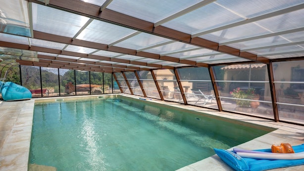 An indoor swimming pool with clear glass walls, a tiled floor, and a roof structure. - Les Gargoulettes (Lauris)