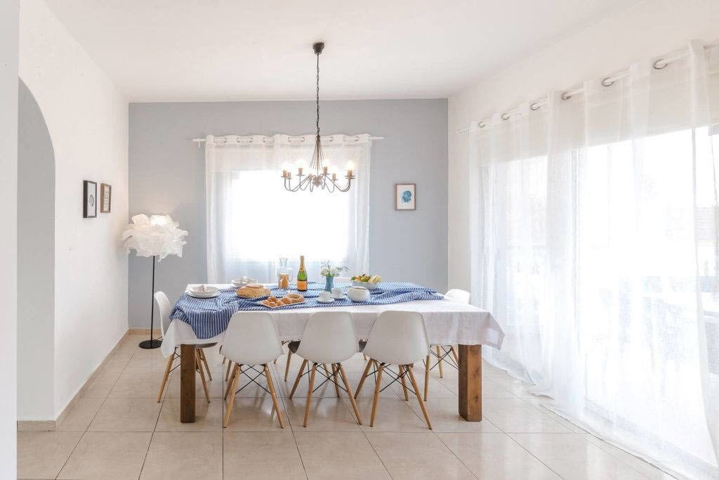 A dining area with a table set for four, a chandelier, and a large window with sheer curtains.