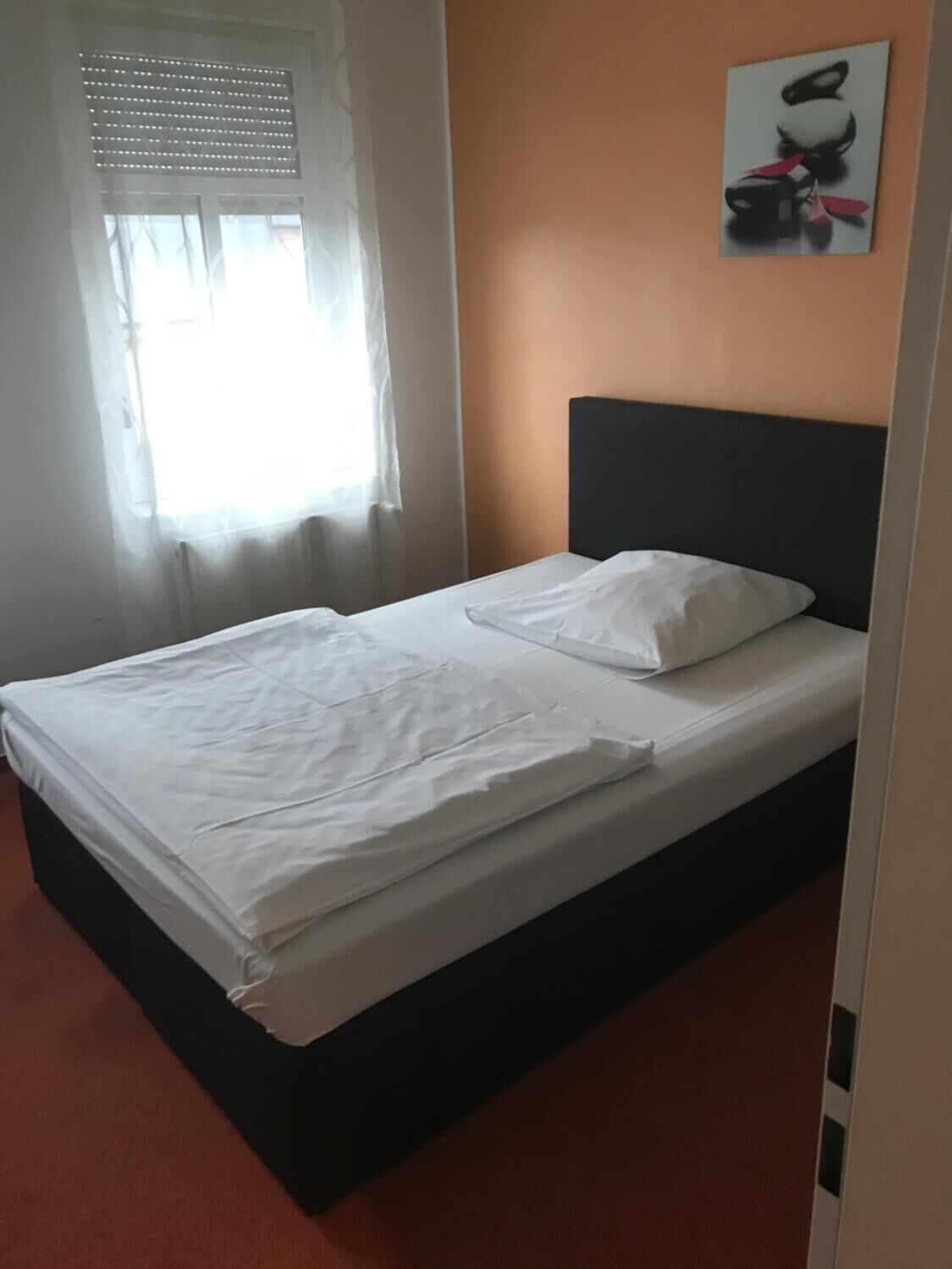 Un lit simple avec du linge de lit blanc, dans une pièce dotée d’une fenêtre et d’un tableau au mur.