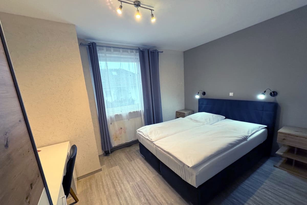A modern hotel room with a large bed, a bedside table, a nightstand, a desk with a chair, and a window with curtains.