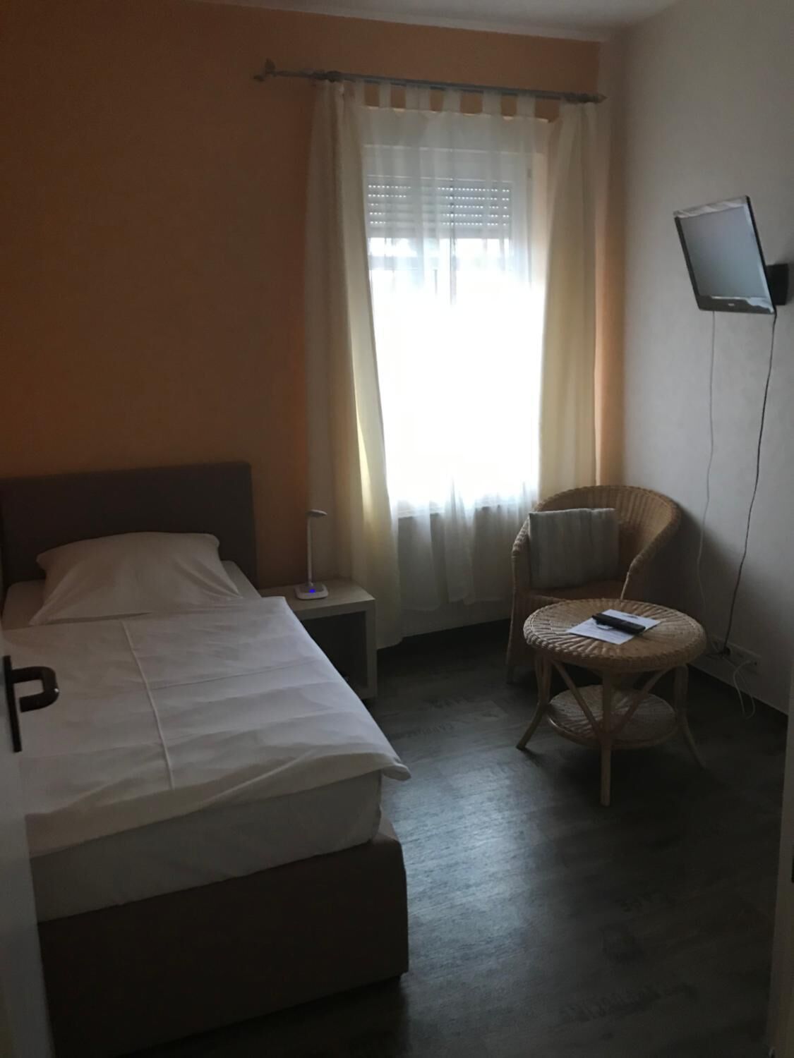 A hotel room with a bed, a bedside table with a lamp, a chair, and a flat-screen TV mounted on the wall.