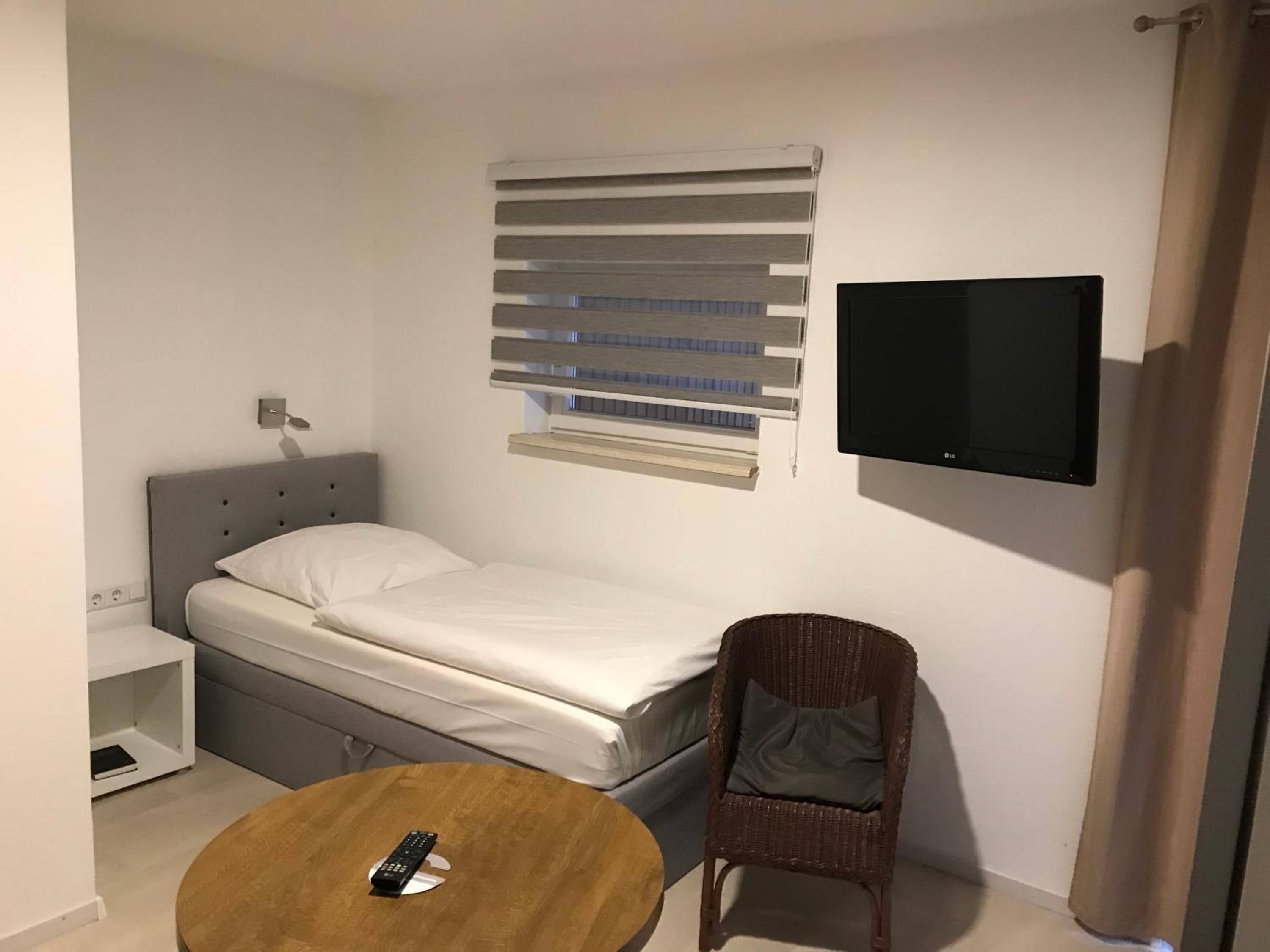Een compacte, moderne kamer met een bed, een aan de muur bevestigde televisie, een raam met zonwering, een klein tafeltje en een stoel.