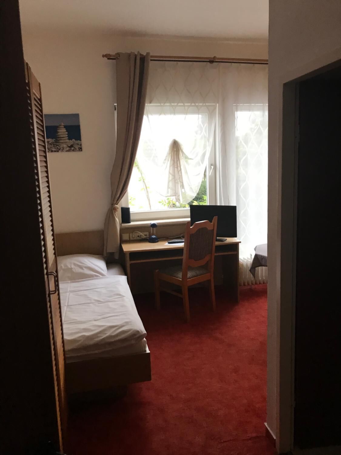 Une chambre avec un lit, un bureau et une chaise. Il y a une fenêtre avec des rideaux, un radiateur et un tableau au mur.