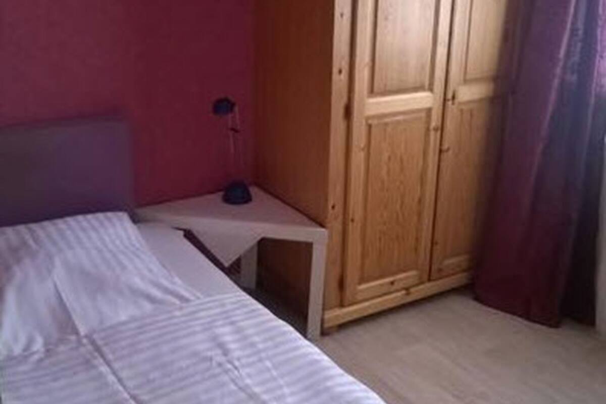 A room with a bed, a small table with a lamp, and a wooden wardrobe.