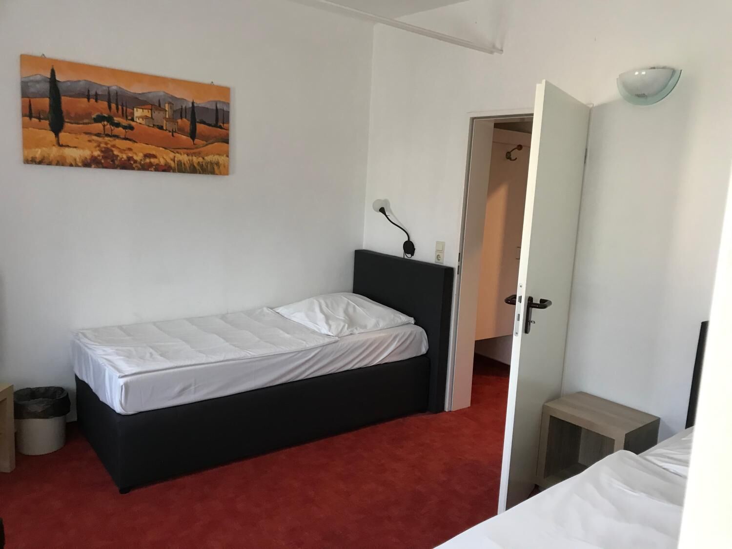 Une petite pièce simple, avec un lit, un tableau accroché au mur et une porte donnant sur une autre pièce.