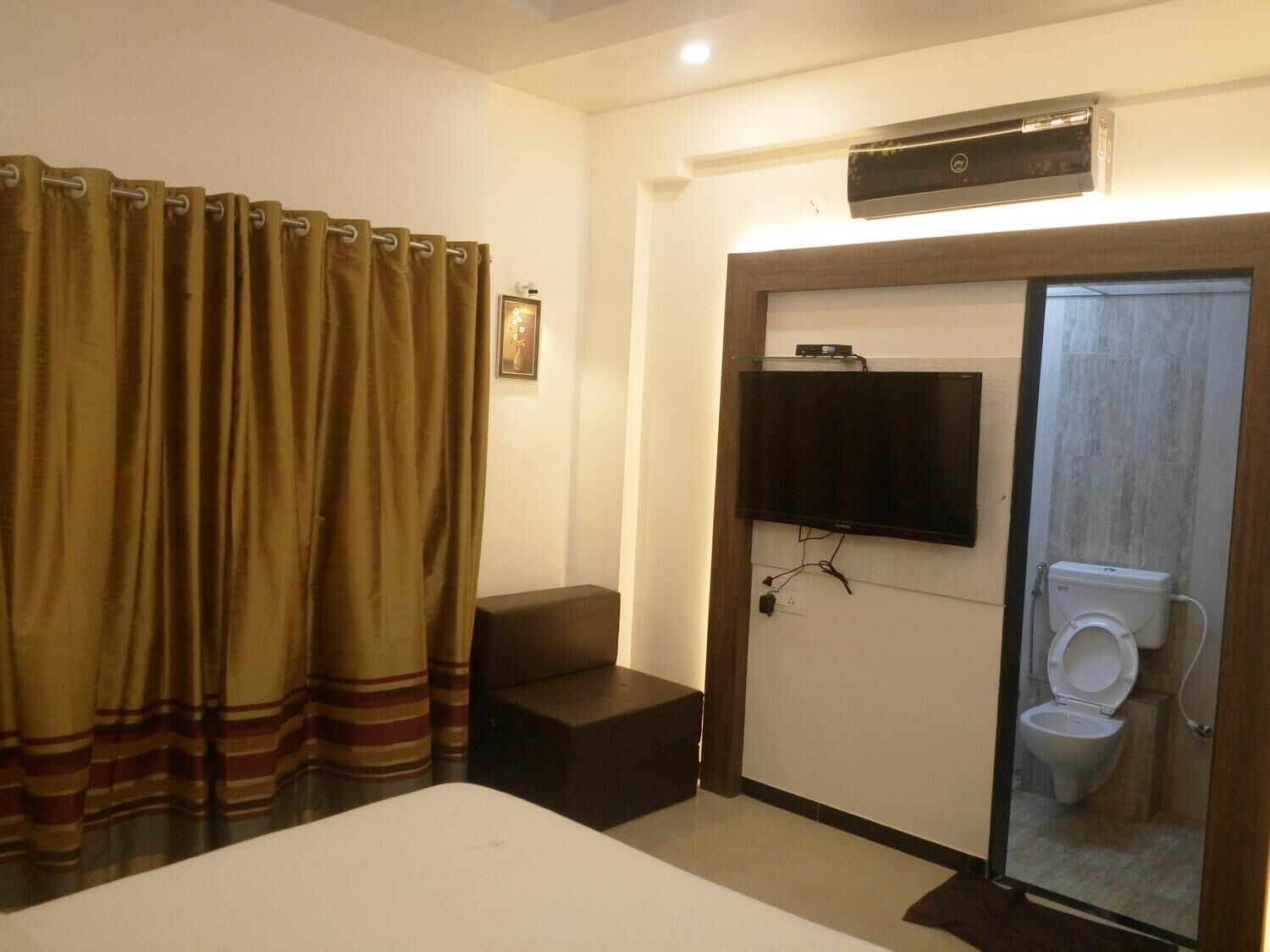 A hotel room with a bed, a television mounted on the wall, a small seating area, and a bathroom with a toilet and shower.