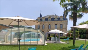 A swimming pool with a glass enclosure, outdoor seating, and a large building in the background. - Hôtel la Tour Perrier (Eyrans)