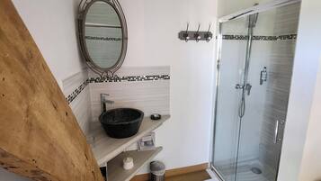 A bathroom with a wooden floor, a round mirror, a black basin, a glass shower enclosure, and a tiled wall.