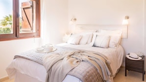 A bedroom with a bed, bedside table, and a window with a view of palm trees. - Villa Isabella (Pegeia)