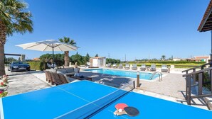 A pool area with lounge chairs, a table, and a car parked nearby. - Villa Isabella (Pegeia)