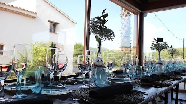 Una mesa de comedor decorada con varias copas de vino y platos, un jarrón con flores y vistas a un edificio blanco y follaje exterior.