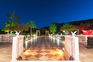 Un pati exterior amb un paviment de pissos quadriculats, balustrades blanques i arbres il·luminats.