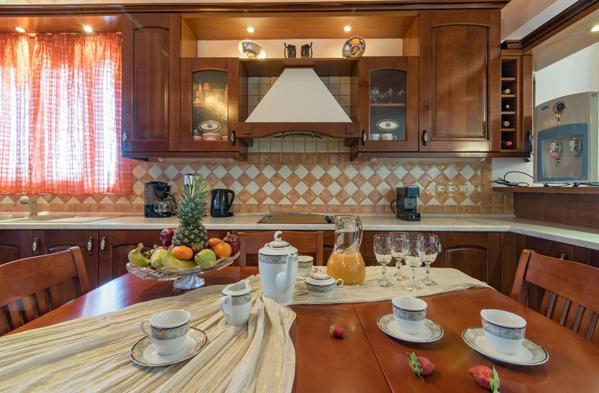 A kitchen with wooden cabinets, a tiled backsplash, and a dining table set for four with cups, saucers, and a fruit bowl.