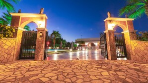 Uma entrada com calçamento de pedra, duas arcadas iluminadas, palmeiras e um edifício ao fundo.
