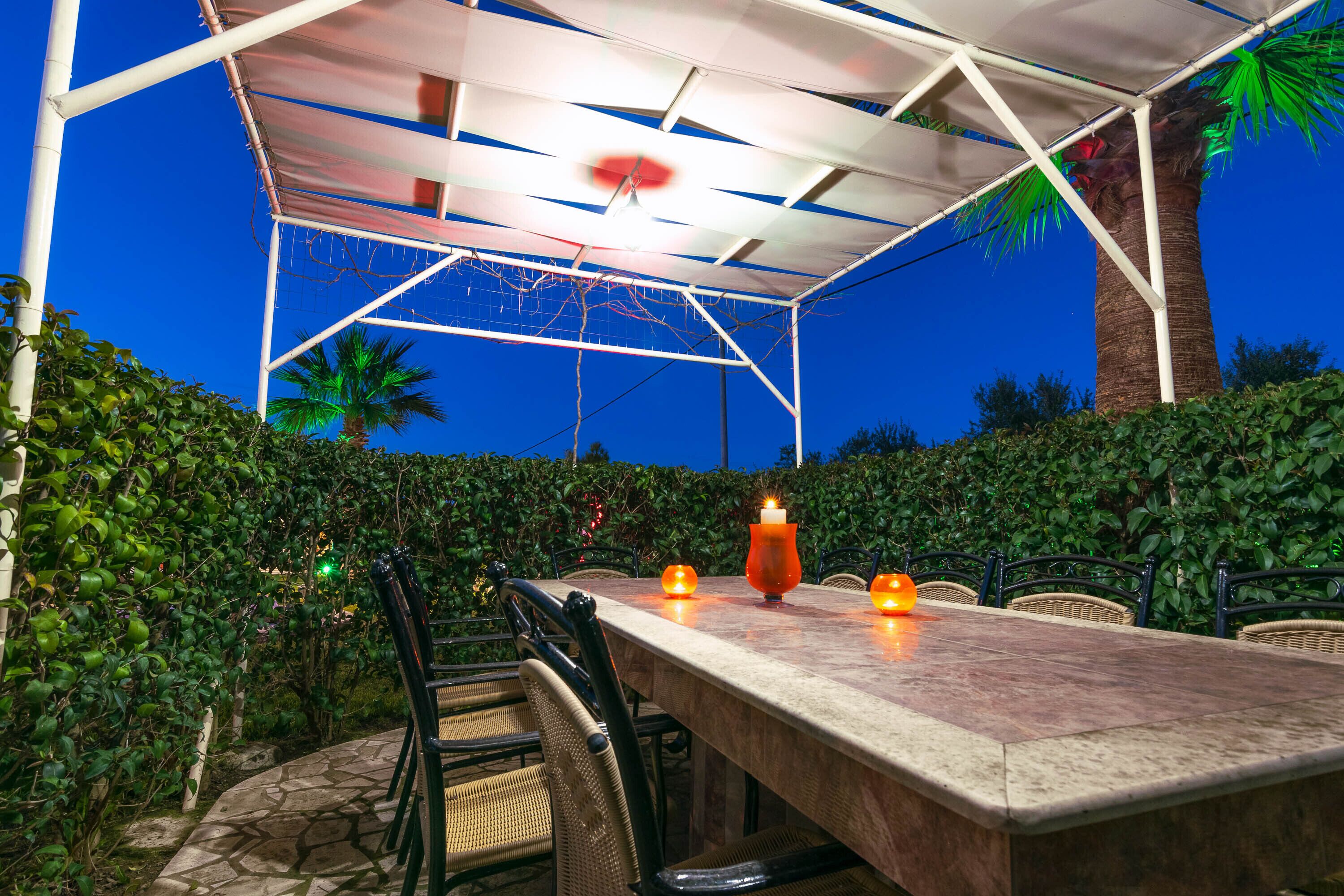 An outdoor dining area with a covered table and chairs, surrounded by lush greenery and a palm tree.