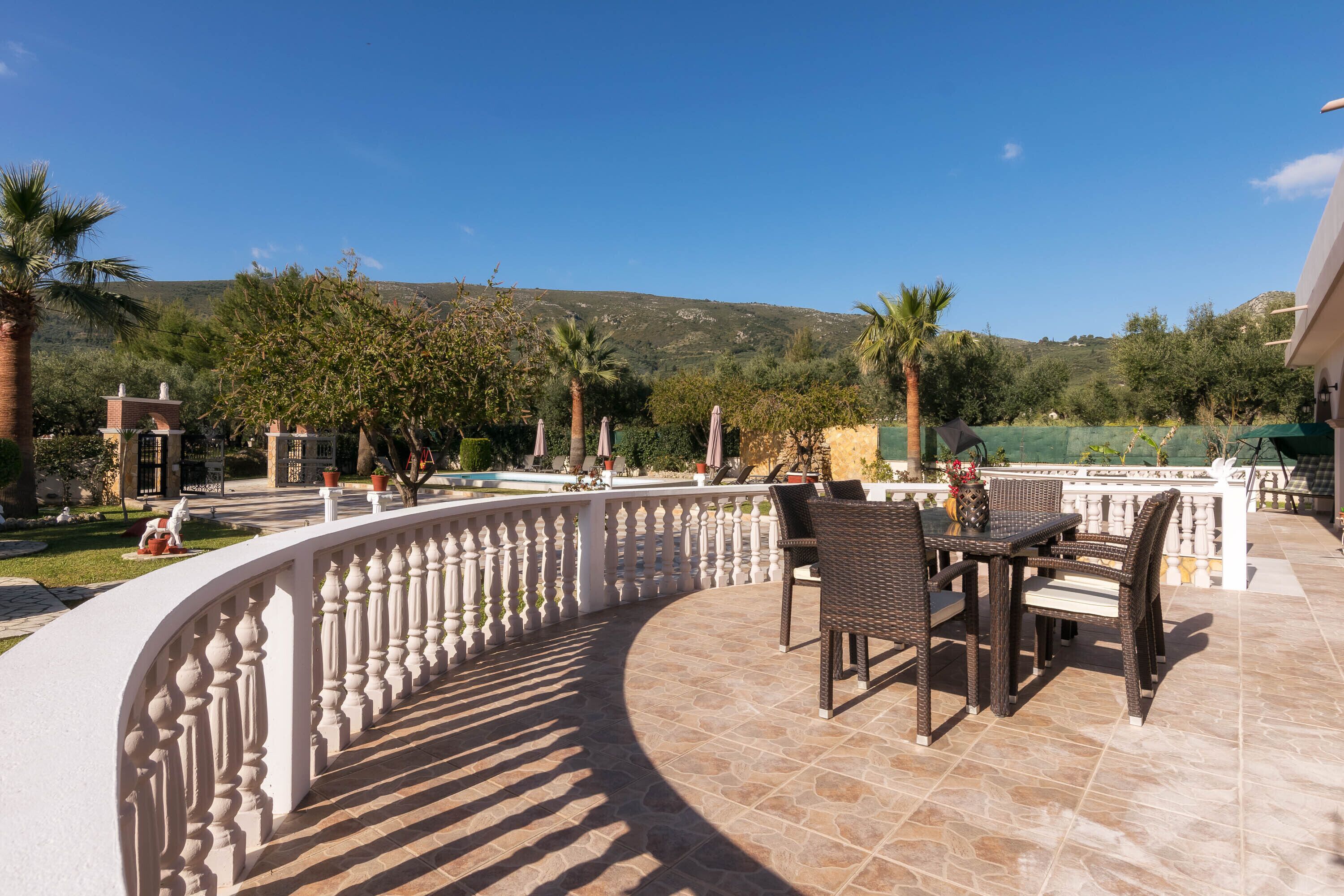 A curved outdoor patio with a white balustrade, wicker furniture, and a view of a pool and greenery.