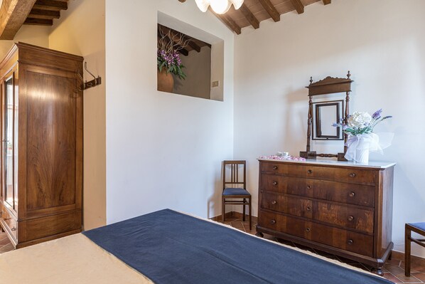 A bedroom with a wooden wardrobe, a dresser, a bed with a blue bedspread, a chair, and a mirror on the dresser. - Il Poderino (Castellina in Chianti)