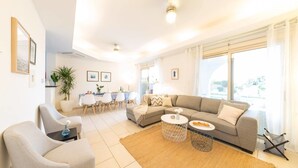A modern living room with a sofa, coffee table, and chairs. There is a dining area with a table and chairs. The room has large windows and a ceiling light. - Villa Jaimi (Pegeia)