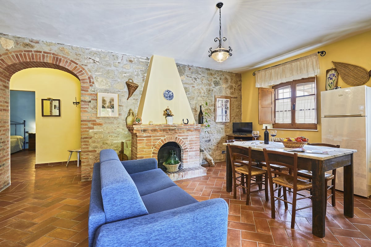 A cozy living room with a blue sofa, a fireplace, a dining area with wooden chairs, and a kitchen with a refrigerator.