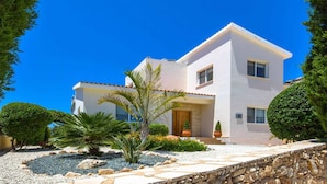 Una casa moderna de dos pisos con fachada blanca, un muro de piedra al frente y un jardín bien cuidado con diversas plantas y una palmera.