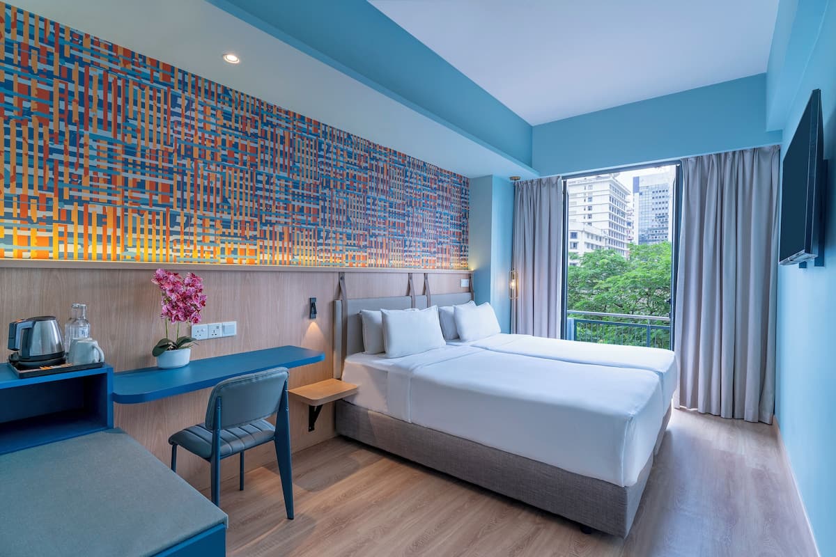 Una habitación de hotel moderna con una cama grande, un escritorio con una silla, una mesita con una planta y vistas a la ciudad a través de un ventanal amplio.