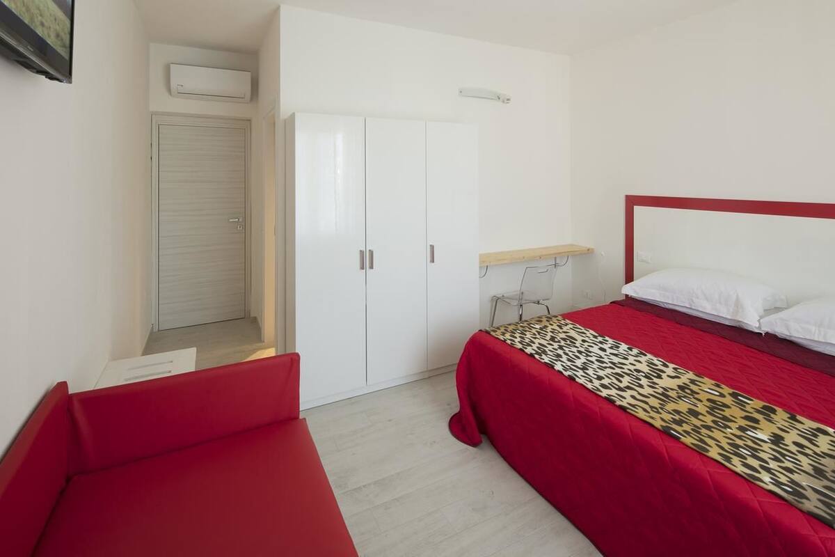 Un dormitorio con un sofá rojo, una cama con edredón estampado, un armario y una mesita.