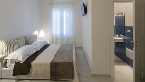 Een slaapkamer met een bed, bedlampen, een raam met dunne gordijnen en een badkamer zichtbaar door een open deur.