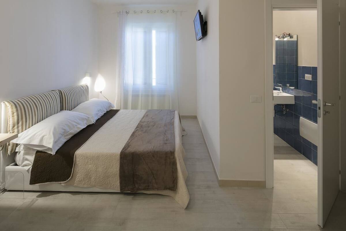 Un dormitorio con cama, lámparas de noche, una ventana con cortinas traslúcidas y un baño visible a través de una puerta abierta.