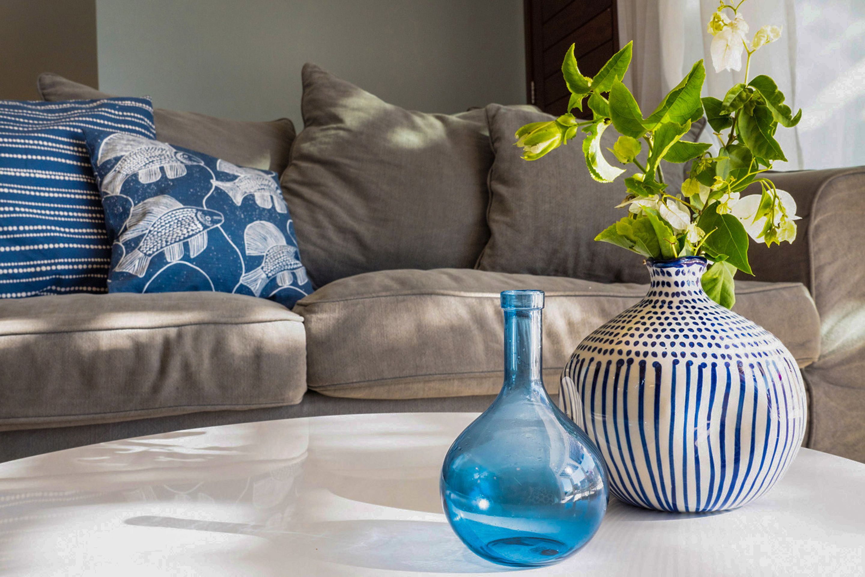 Un salón con un sofá beige, un florero azul con plantas verdes y una botella de vidrio azul sobre una mesa blanca.