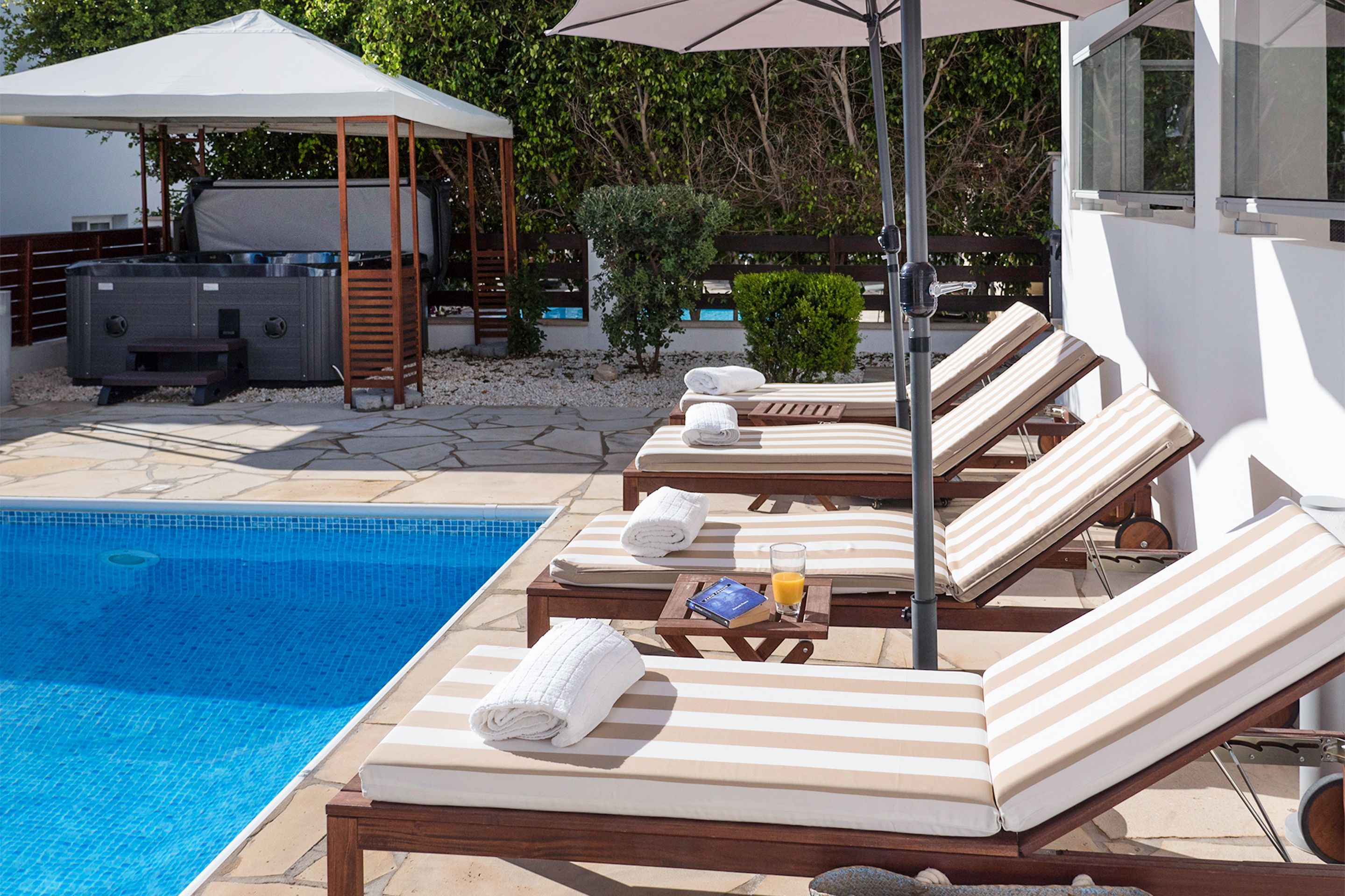 Uma área à beira da piscina com espreguiçadeiras, uma banheira de hidromassagem e um espaço coberto para sentar.