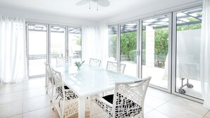 Un vaste espace dédié à la salle à manger, avec une table en verre et des chaises en osier blanc, de grandes fenêtres avec des rideaux et un ventilateur de plafond.
