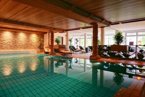 An indoor swimming pool with tiled flooring, wooden ceiling beams, and lounge chairs along the poolside. - Landhaus Haake (Bad Zwischenahn)
