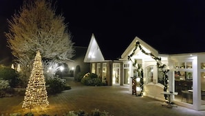 Un hotel con un árbol cubierto de nieve y un árbol de Navidad, ambos iluminados. Se observa un edificio con techo triangular y un patio cubierto con una mesa y sillas.