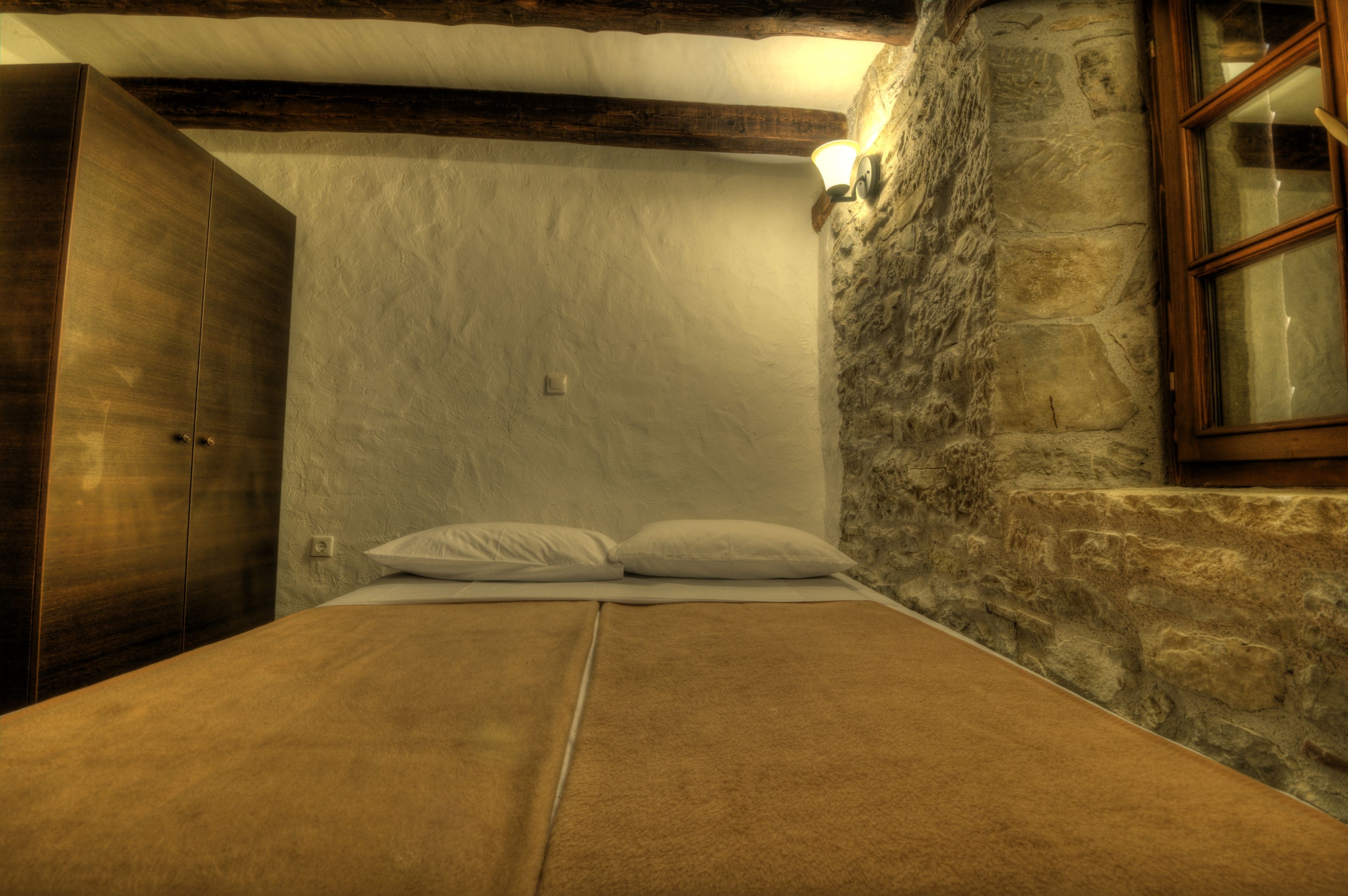 A bedroom with a bed, wooden wardrobe, stone wall, and a window.