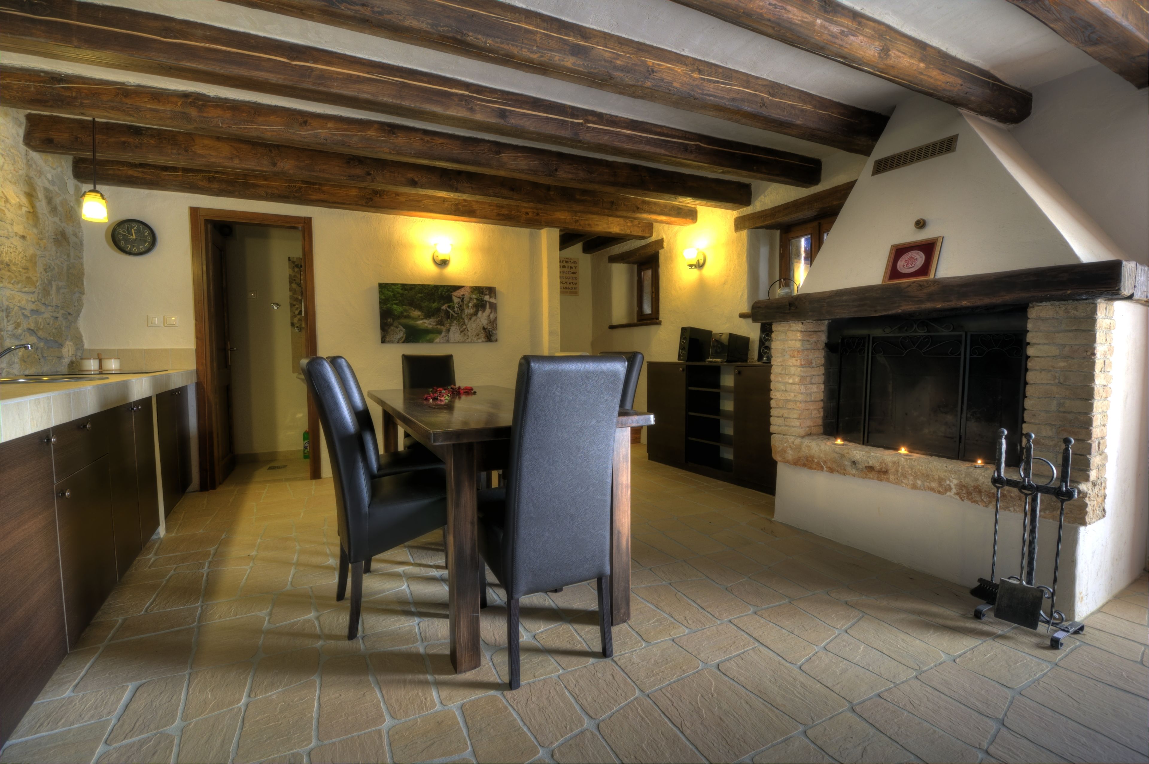 A dining area with a stone floor, wooden beams, a fireplace, and a kitchen visible in the background.